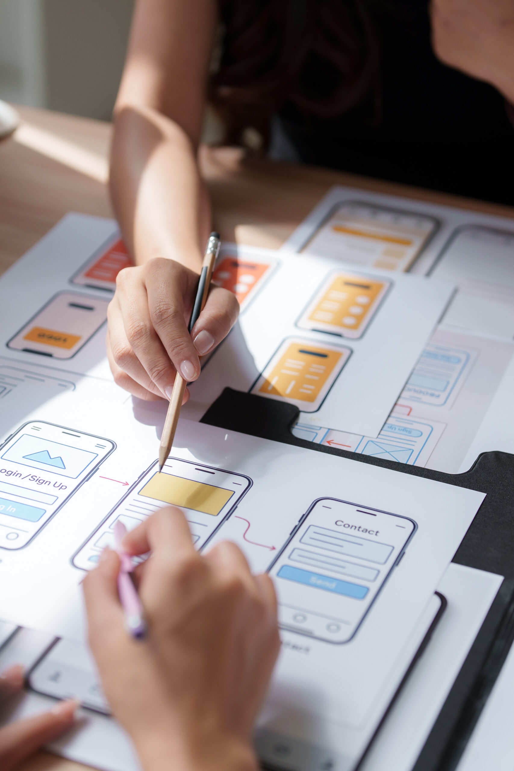 Hands working with pencils on papers displaying various mobile app screen designs and user interfaces